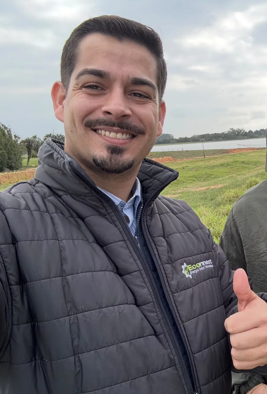 Juliano Lamas sorrindo e fazendo sinal de positivo em uma propriedade com lago ao fundo