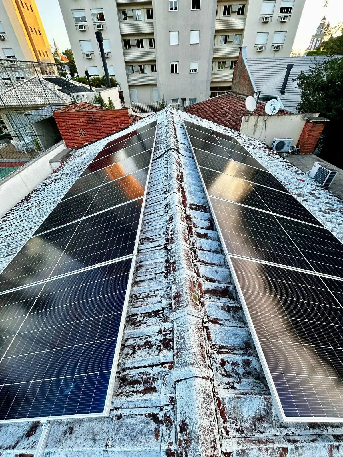 Módulos fotovoltaicos na cumeeira de um telhado, com prédios ao fundo