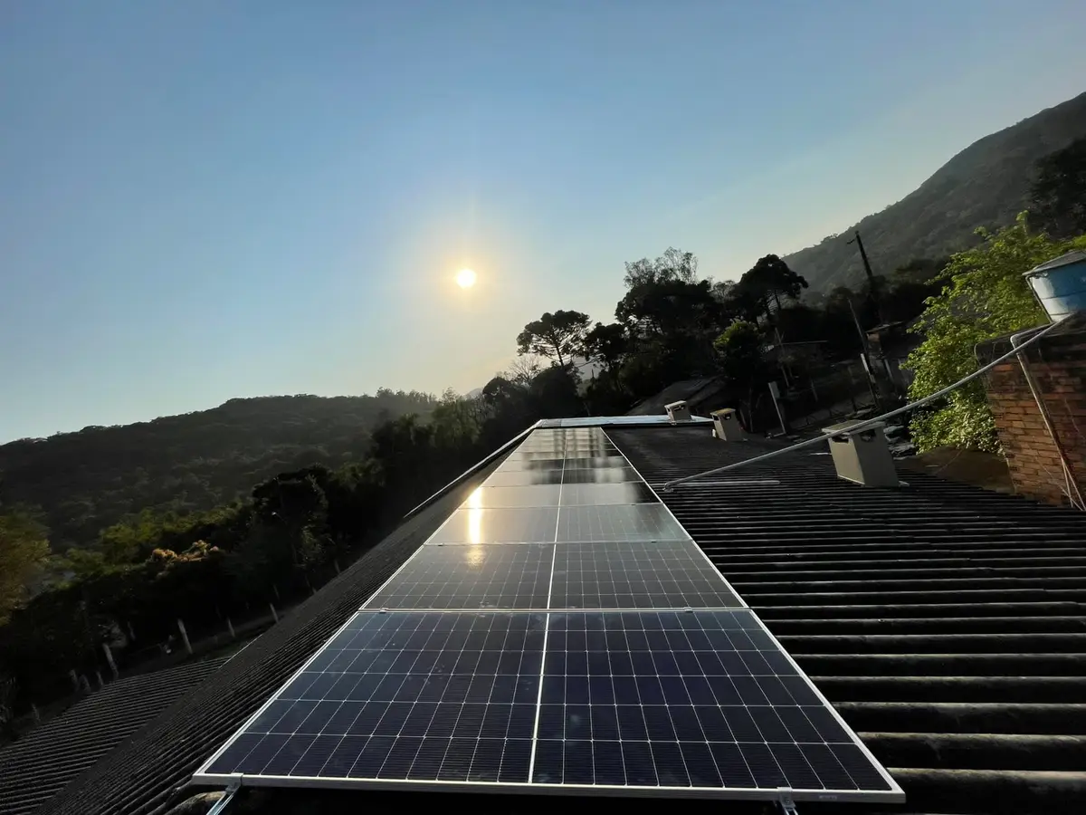 Módulos fotovoltaicos em telhado ao entardecer, com morros ao fundo